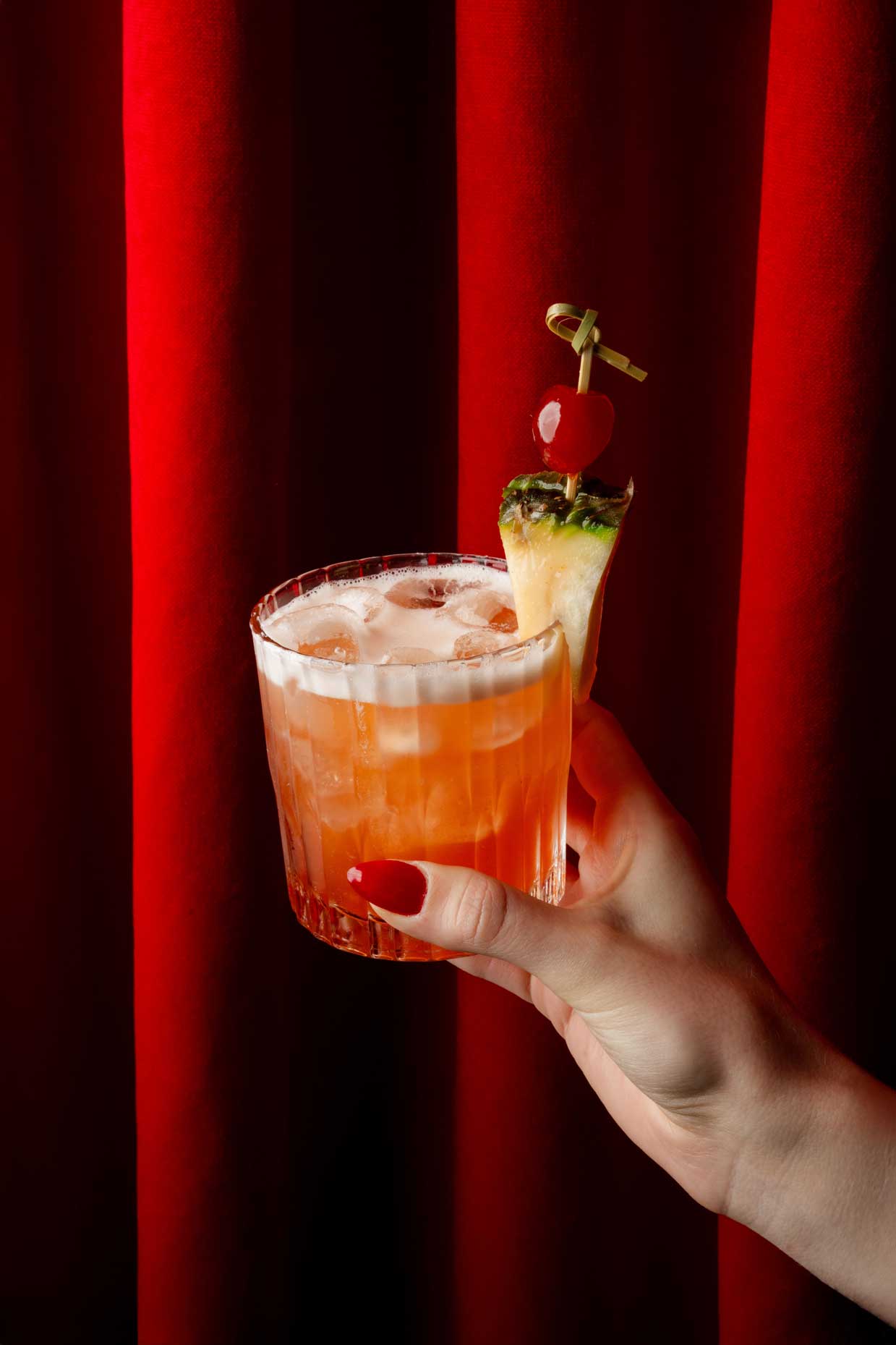 Hand holding a vibrant cocktail with a garnish and red curtain background.